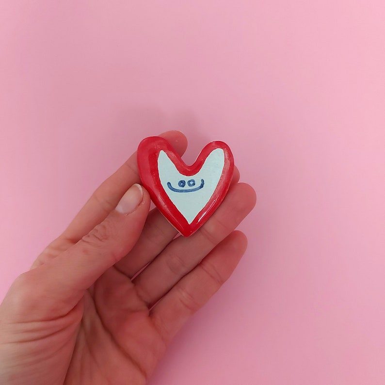 May include: A red heart-shaped ceramic ornament with a white face and blue eyes and a smile. The ornament is being held in a hand.