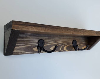 May include: A wooden shelf with two black metal hooks. The shelf is stained a dark brown color and has a slightly angled front edge. The hooks are attached to the bottom of the shelf and are angled slightly upwards.