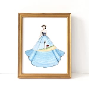 Puede incluir: Una impresión en acuarela enmarcada que presenta a una mujer con un vestido azul con una escena de faro. El vestido tiene un corpiño a rayas y una falda fluida que representa una playa y el océano. El marco es dorado.