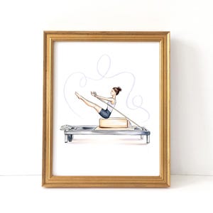 Könnte beinhalten: Ein gerahmter Aquarelldruck zeigt eine Person, die Pilates auf einem Reformer-Gerät macht. Das Kunstwerk hat einen goldenen Rahmen, einen hellblauen Hintergrund und eine Person in einem weißen Oberteil und dunklen Shorts. Der Pilates-Reformer ist silber und beige.