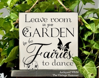 May include: White wooden sign with black text that reads "Leave room in your GARDEN for the fairies to dance". The sign has a distressed look and a butterfly graphic. The sign is hanging from a twine rope.