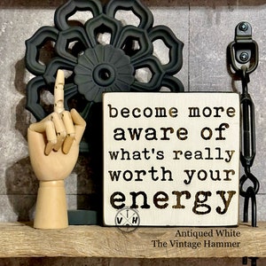 May include: White wooden sign with black text that reads "become more aware of what's really worth your energy". The sign has a brown distressed wood background and a black and white logo with the letters "VH" in the bottom left corner. The sign is on a wooden surface with a black metal flower design and a black metal hook in the background.