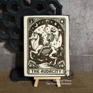 May include: A wooden sign with a black and white illustration of a skeleton riding a unicorn. The sign reads "The Audacity" and is on a small easel. The sign is distressed and has a white background.