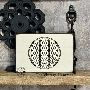 May include: A white wooden sign with a black and white geometric design, known as the Flower of Life, in the center. The sign is hanging from a black metal hook and has the text "The Vintage Hammer" at the bottom.