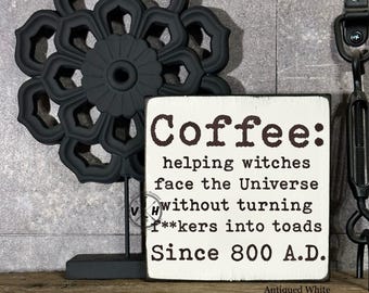 May include: A white wooden sign with black text that reads "Coffee: helping witches face the Universe without turning f**kers into toads Since 800 A.D.". The sign is sitting on a wooden surface with a black metal flower decoration behind it.