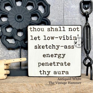 May include: White wooden sign with black text that reads "thou shall not let low-vibin' sketchy-ass energy penetrate thy aura". The sign is distressed and has a vintage look. The text is in a typewriter font. The sign is on a wooden surface.