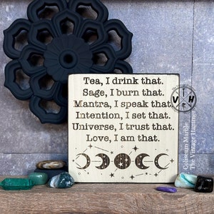 May include: A wooden sign with a white background and black text. The sign reads "Tea, I drink that. Sage, I burn that. Mantra, I speak that. Intention, I set that. Universe, I trust that. Love, I am that." The sign also has a moon and star design.