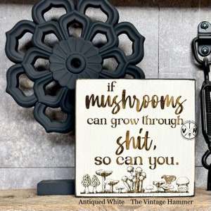 May include: A white wooden sign with the text "if mushrooms can grow through shit, so can you." in gold lettering. The sign is decorated with a row of mushrooms at the bottom and a small logo in the top right corner. The sign is sitting on a black surface.