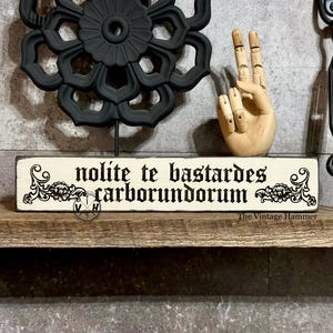 May include: A wooden sign with the phrase "nolite te bastardes carborundorum" in black gothic font. The sign is cream-colored with decorative floral accents. A black metal floral sculpture and a wooden hand are also visible.