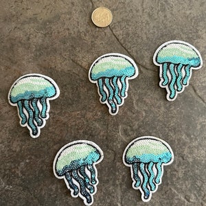 May include: Five iridescent blue and green jellyfish iron-on patches with white outlines. The patches are made of sequins and are perfect for adding a touch of whimsy to clothing, bags, or other accessories.