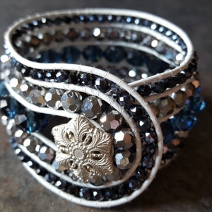Puede incluir: Un brazalete de cuero blanco con múltiples filas de cuentas azules, negras y plateadas. Un dije plateado en forma de copo de nieve está en el centro del brazalete.