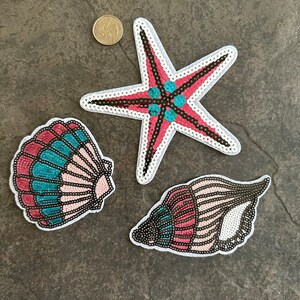May include: Three iron-on patches featuring a pink and blue seashell, a pink and blue seashell with a brown swirl, and a pink and blue starfish with a brown center.