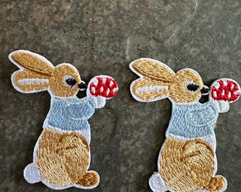 May include: Two embroidered iron-on patches of a brown rabbit holding a red mushroom. The rabbit is wearing a blue shirt.