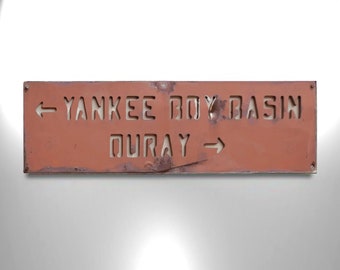 May include: A rusty orange metal sign with white lettering pointing to Yankee Boy Basin and Ouray. The sign has an arrow pointing left for Yankee Boy Basin and an arrow pointing right for Ouray.