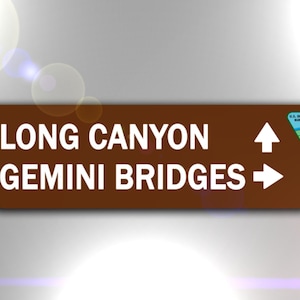 May include: A brown sign with white text that reads "Long Canyon" and "Gemini Bridges". The sign has an arrow pointing up for Long Canyon and an arrow pointing right for Gemini Bridges. The sign also has a green triangle with a river and a pine tree, with the text "U.S. Department of the Interior Bureau of Land Management".