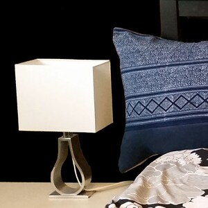 Puede incluir: Una pantalla de l&aacute;mpara cuadrada blanca se asienta sobre una base de l&aacute;mpara plateada con un dise&ntilde;o curvo. Una almohada azul estampada descansa sobre una cama con un edred&oacute;n con estampado floral. El fondo es negro.