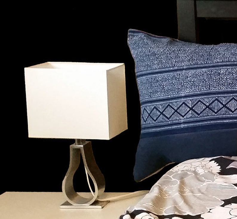 Puede incluir: Una pantalla de l&aacute;mpara cuadrada blanca se asienta sobre una base de l&aacute;mpara plateada con un dise&ntilde;o curvo. Una almohada azul estampada descansa sobre una cama con un edred&oacute;n con estampado floral. El fondo es negro.