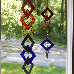 May include: A stained glass suncatcher with a rainbow of colors, including red, orange, yellow, green, blue, and purple. The suncatcher is made of multiple diamond-shaped pieces of glass that are connected together. The suncatcher is hanging in a window.