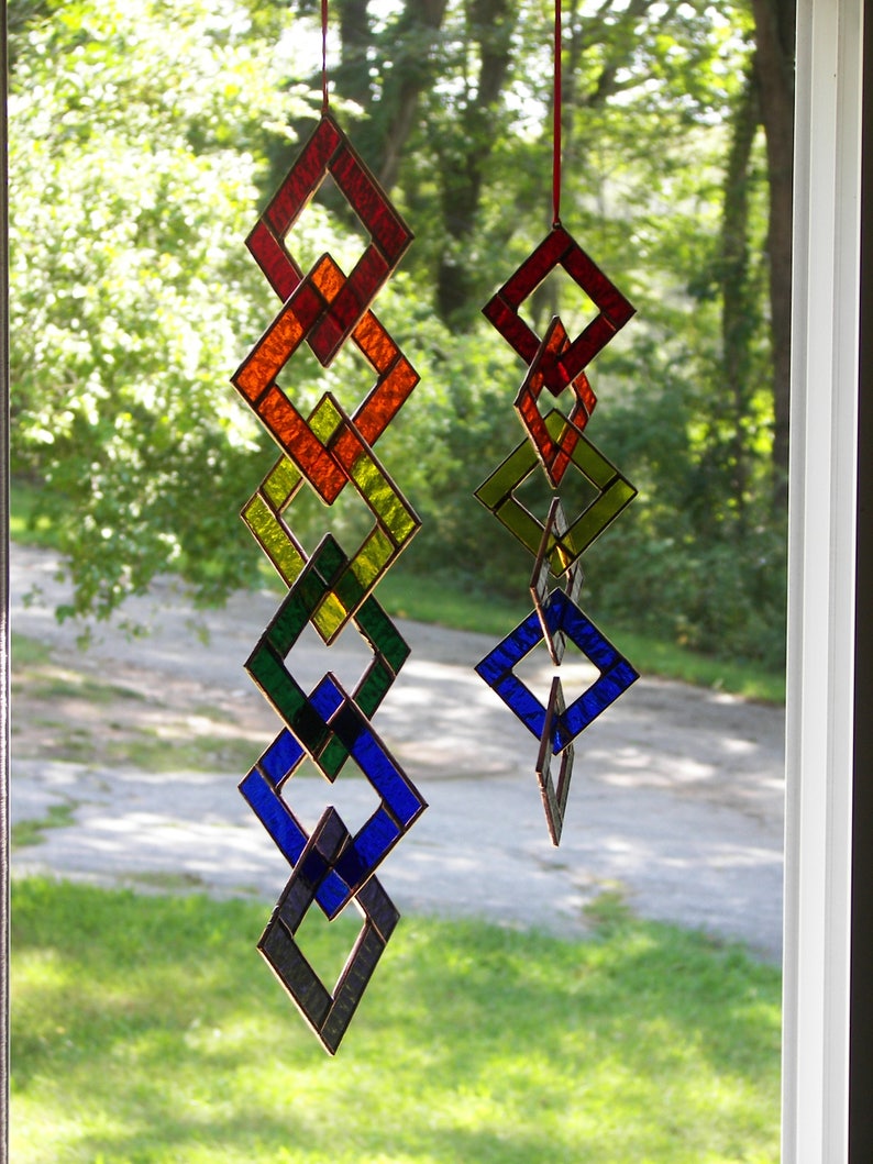 May include: A stained glass suncatcher with a rainbow of colors, including red, orange, yellow, green, blue, and purple. The suncatcher is made of multiple diamond-shaped pieces of glass that are connected together. The suncatcher is hanging in a window.
