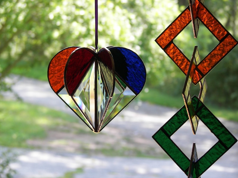 May include: A stained glass heart-shaped ornament with red, blue, and clear glass panels. The ornament is hanging from a string and is in front of a blurred background of green trees.
