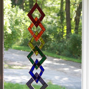 May include: A stained glass suncatcher with a rainbow of colors, including red, orange, yellow, green, blue, and purple. The suncatcher is made of interlocking diamond shapes and hangs from a red string.