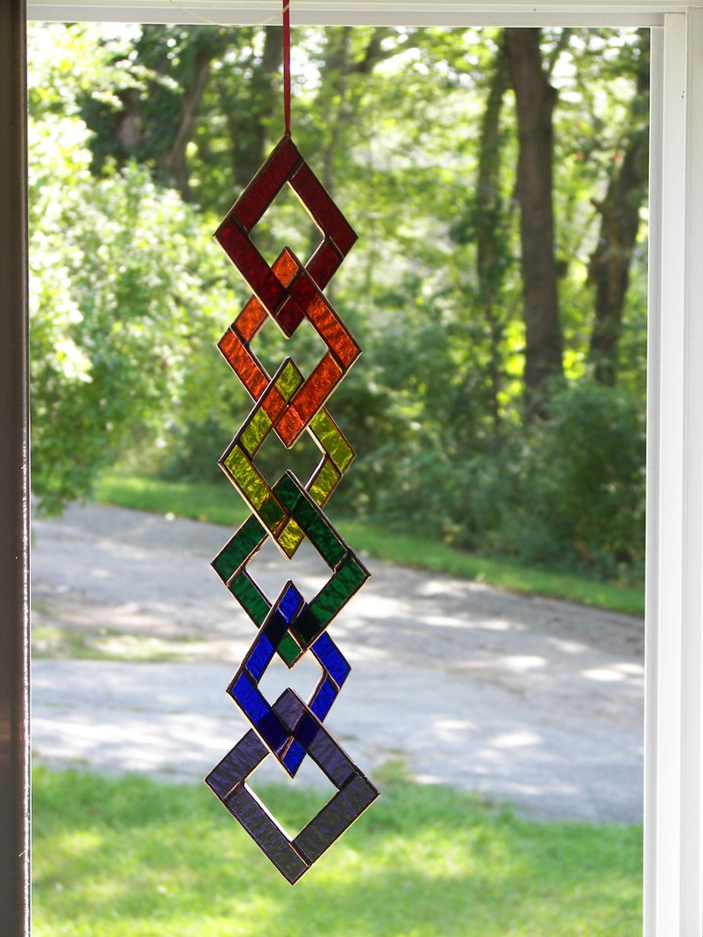 May include: A stained glass suncatcher with a rainbow of colors, including red, orange, yellow, green, blue, and purple. The suncatcher is made of interlocking diamond shapes and hangs from a red string.