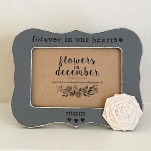 May include: Gray wooden picture frame with a white flower and the text "forever in our hearts" and "flowers in december" with the word "mom" at the bottom.
