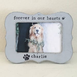 May include: A gray wooden picture frame with a white border and the text "forever in our hearts" and a paw print with the name "charlie" below the picture. The picture shows a golden retriever dog.