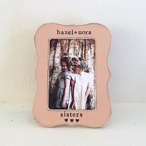 May include: A pink picture frame with a white border and a scalloped edge. The frame has a photo of two girls with the text "hazel & nora sisters" printed on the frame.