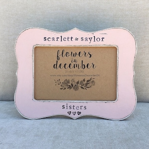 Puede incluir: Un marco de fotos de madera rosa con un acabado blanco desgastado. El marco tiene un borde festoneado y presenta el texto "scarlett & saylor flowers in december design studio www.etsy.com/shop/FlowersInDecemberDS sisters" con tres corazones.
