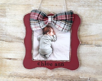 May include: A red framed photo holder with a plaid bow and a photo of a baby. The frame has the name "chloe ann" printed on it. The bow is red, white, and black plaid. The frame is hanging by a string.
