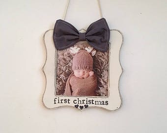 May include: A framed photo with a baby wearing a knitted hat and wrapped in a blanket. The frame is off-white with a dark gray bow at the top and the words "first christmas" at the bottom.