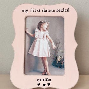 May include: A pink wooden picture frame with the text "my first dance recital" at the top and "emma" with three hearts at the bottom. The frame holds a photo of a young girl in a pink dress.