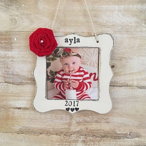 Puede incluir: Adorno de marco de fotos de madera blanca con una flor de tela roja, una foto de una ni&ntilde;a peque&ntilde;a y el texto "ayla 2017" con tres corazones negros.
