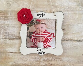 May include: White wooden picture frame ornament with a red fabric flower, a photo of a baby girl, and the text "ayla 2017" with three black hearts.