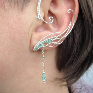 May include: Silver ear cuff with a delicate, wing-like design and a dangling chain of light green crystals.