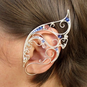 May include: A silver wire ear cuff with blue and clear crystals. The cuff is designed to wrap around the ear and has a delicate, intricate design.