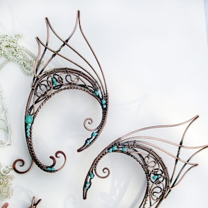May include: A pair of copper wire earrings with turquoise beads, shaped like wings. The earrings are delicate and intricate, with a whimsical design.