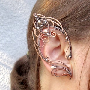 May include: A copper wire ear cuff with a delicate, intricate design and small white pearl accents. The cuff is designed to wrap around the ear and is a unique and stylish accessory.