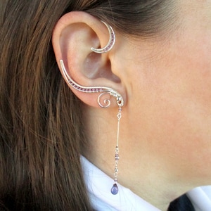 May include: A silver ear cuff with purple beads wraps around the upper ear, and a dangling earring with a purple stone hangs from the lower earlobe.