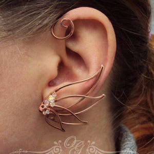 May include: A copper ear cuff with a delicate, intricate design featuring multiple curved wires and small clear crystals. The cuff is worn on the upper part of the ear, with a separate hoop-shaped piece worn lower down.