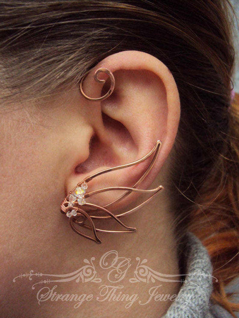May include: A copper ear cuff with a delicate, intricate design featuring multiple curved wires and small clear crystals. The cuff is worn on the upper part of the ear, with a separate hoop-shaped piece worn lower down.