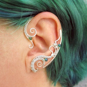 May include: A silver ear cuff with green beads, designed to wrap around the ear. The cuff is made of wire and has a delicate, intricate design.