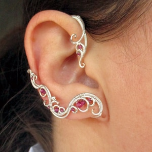 May include: A silver ear cuff with a delicate, intricate design featuring a series of small, pink beads. The cuff is designed to wrap around the upper part of the ear.