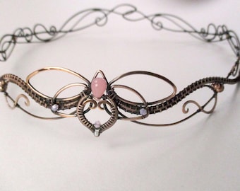 May include: A delicate copper wire crown with a pink gemstone and smaller white gemstones. The crown is made of wire that is woven together to create a floral design.