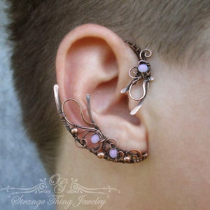 May include: A copper-colored ear cuff with a delicate, wire-wrapped design. The cuff features small, light purple beads and intricate swirls. The ear cuff is designed to wrap around the ear, creating a unique and artistic accessory.
