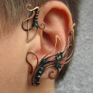 May include: A copper wire ear cuff with a delicate, intricate design. The cuff features a series of loops and swirls, accented with small, emerald green crystals. The cuff is designed to wrap around the upper part of the ear.