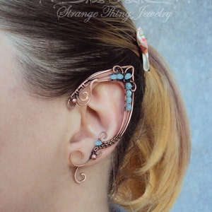 May include: A copper ear cuff with a delicate, intricate design. The cuff is adorned with small, light blue gemstones. The cuff is worn on the upper part of the ear, extending up and around the ear.