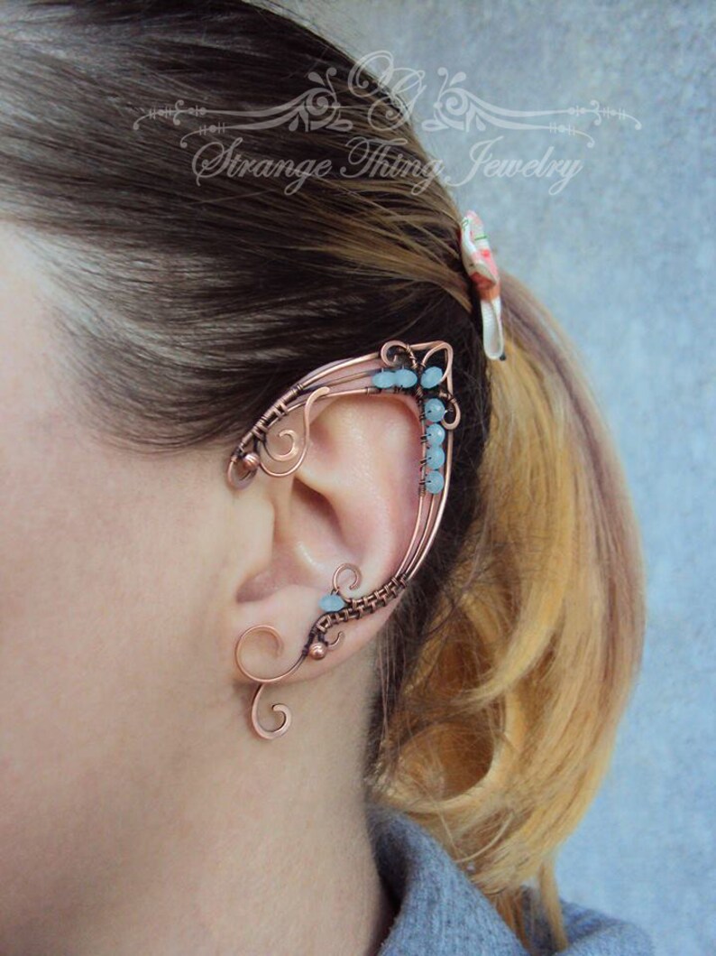 May include: A copper ear cuff with a delicate, intricate design. The cuff is adorned with small, light blue gemstones. The cuff is worn on the upper part of the ear, extending up and around the ear.