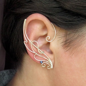 May include: Silver ear cuff with a delicate, intricate design featuring a small blue bead. The cuff is designed to wrap around the upper ear.